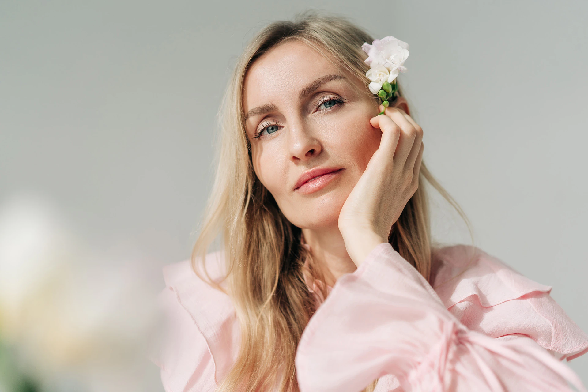 Eine Frau mit langen blonden Haaren und rosa Bluse stützt den Kopf in die Hand und trägt eine weisse Blüte hinter dem Ohr.
