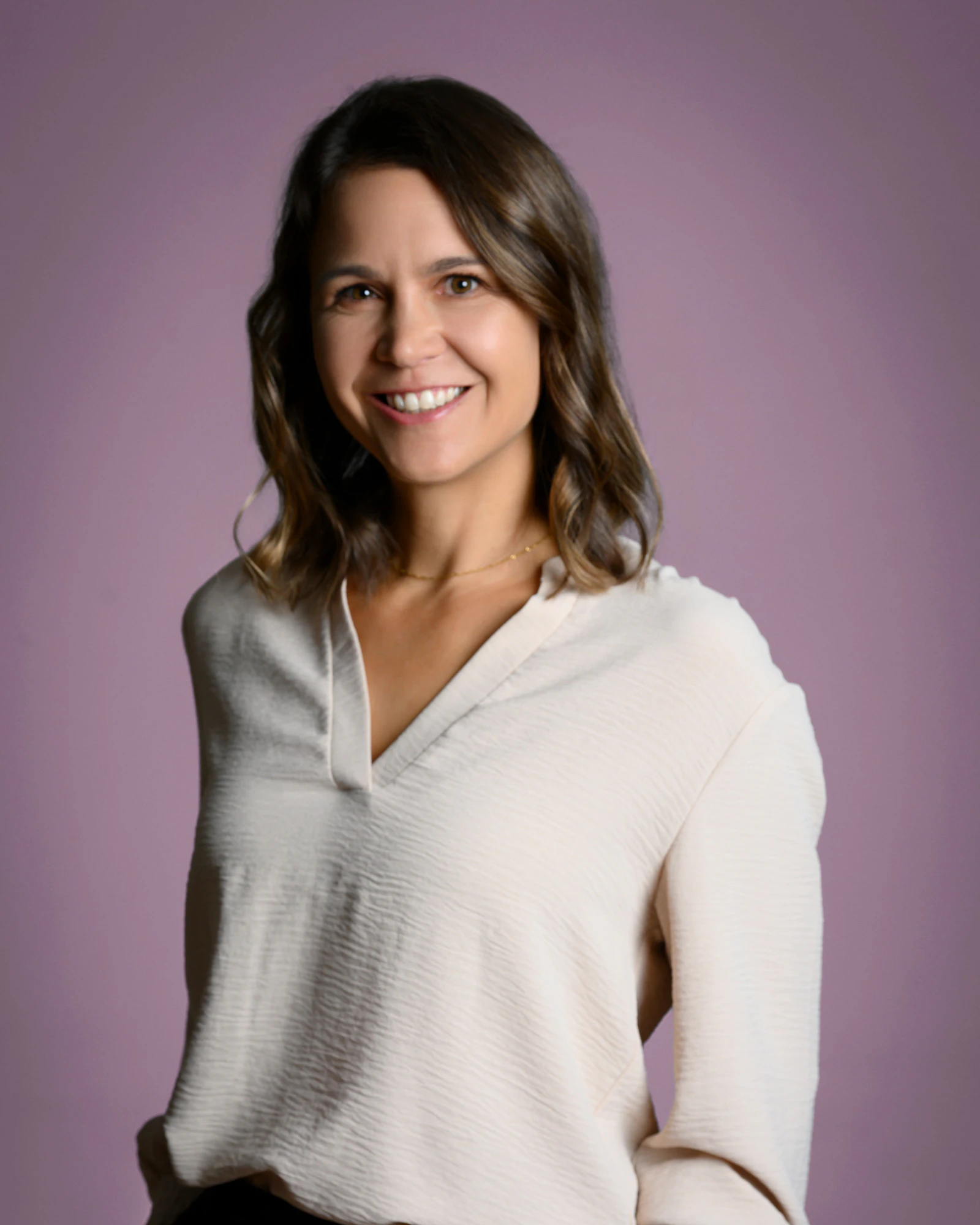 Porträt von Sara Rohrer: eine lächelnde Frau mit schulterlangen braunen Haaren und heller Bluse vor violettem Hintergrund.